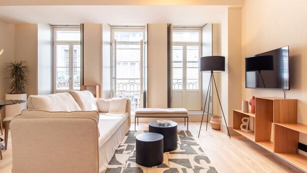 A modern living room with a sofa, coffee table, and a TV. There are large windows, a floor lamp, and a bookshelf. - Catarina 392 | Chic Studio by LovelyStay (Porto)