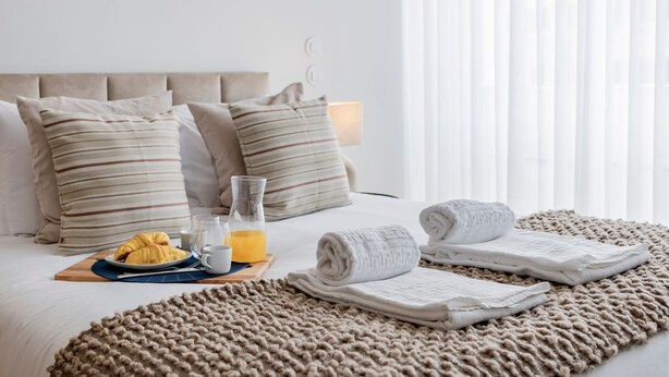 Ein ordentlich bezogenes Bett mit Kissen, ein Glas Orangensaft, ein Croissant und ein aufgerolltes Handtuch auf einem strukturierten Teppich.