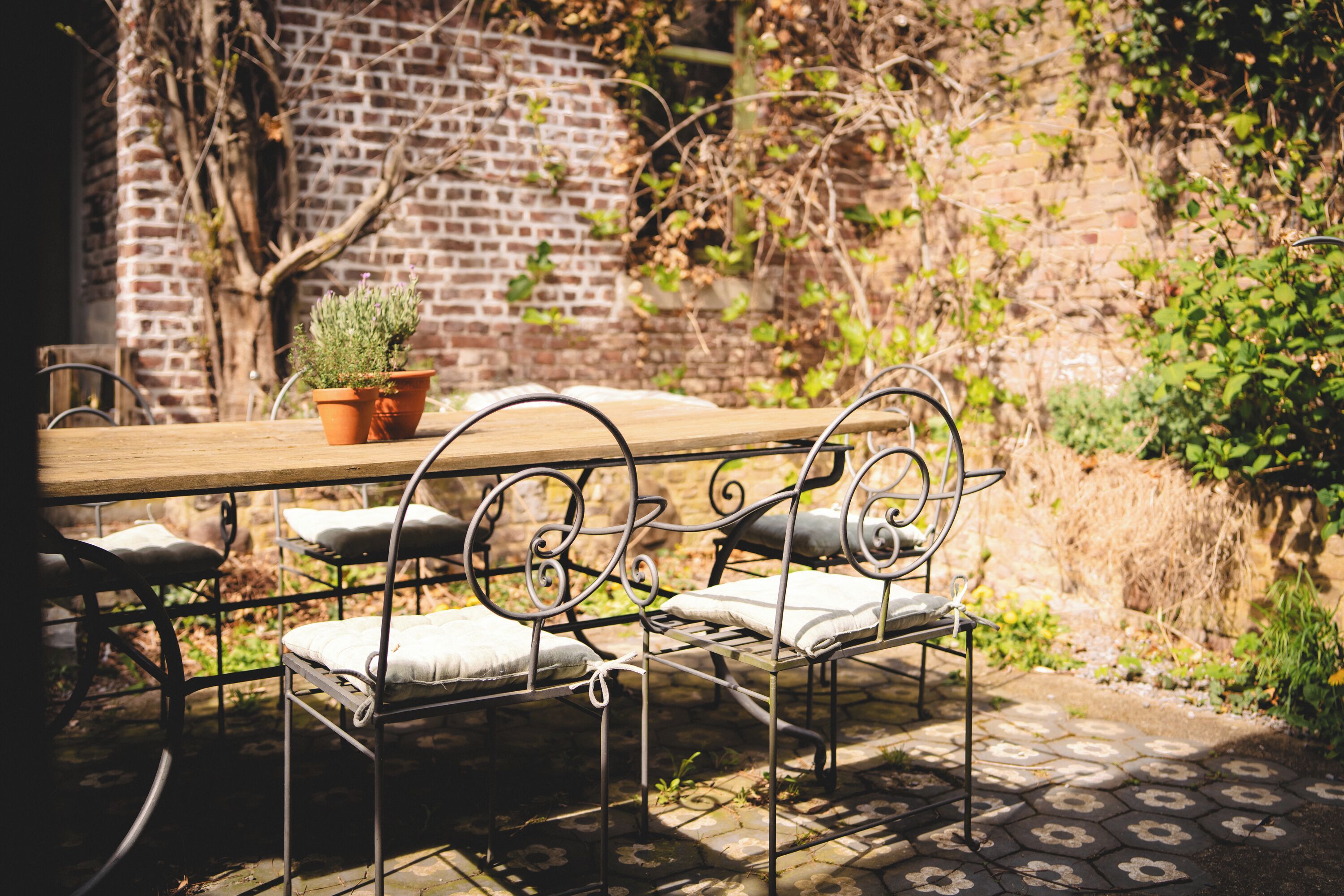 A rustic outdoor dining area with a wooden table and metal chairs, surrounded by a brick wall and greenery.