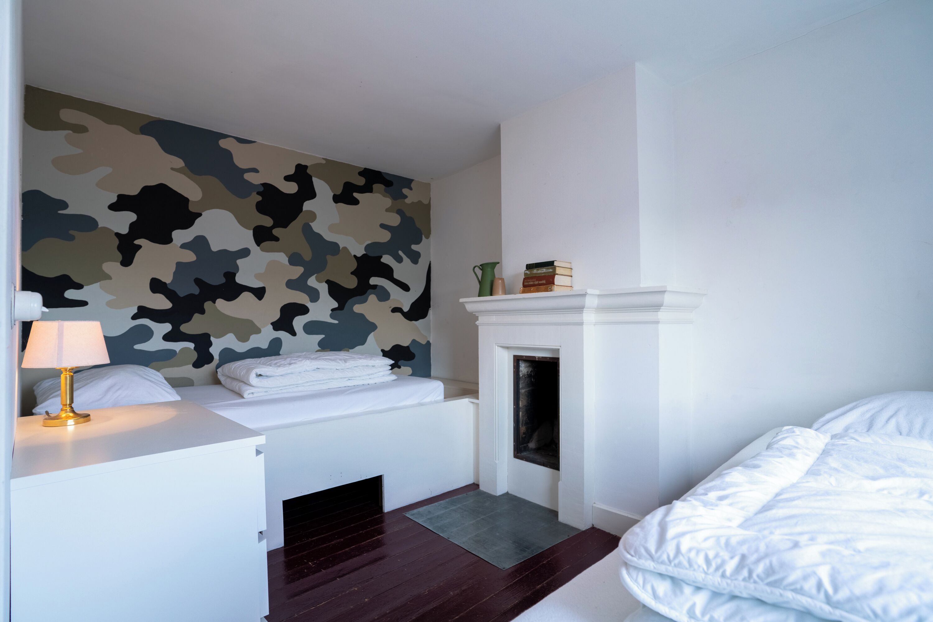 Un dormitorio con una pared estampada en camuflaje, una cama blanca, una chimenea pequeña y una mesita de noche con una lámpara.