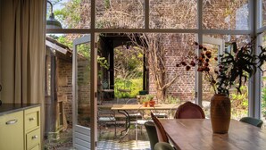 Un comedor con mesa y sillas de madera, un jarrón con flores, y vista a una pared de ladrillo y follaje exterior.