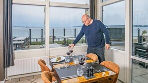 Un comedor con una mesa dispuesta para dos personas, ventanas amplias con vista al mar, y un hombre sirviendo vino.