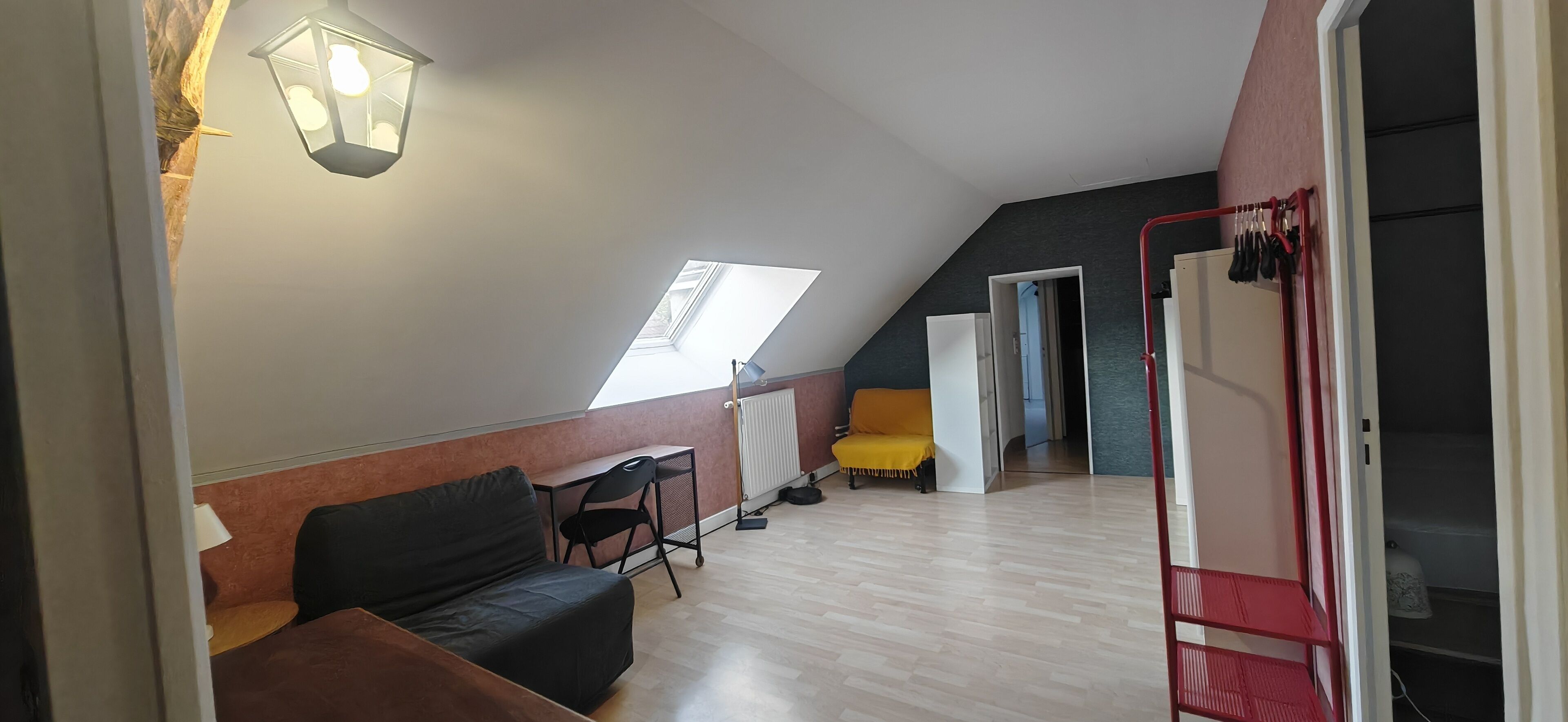 Een loft-achtige kamer met een schuine plafond, een zwarte bank, een klein bureau, een gele stoel en een rode metalen rek.