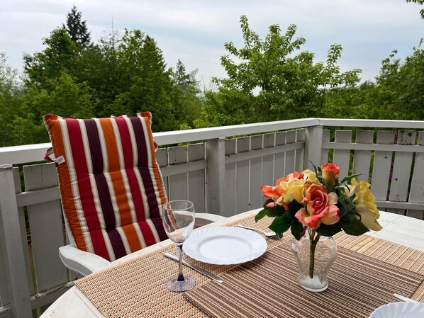 Une table à manger dressée avec une assiette blanche, un verre à vin et un vase rempli de fleurs, le tout placé sur une terrasse dotée d’une balustrade blanche et d’un coussin à rayures.