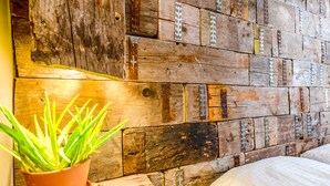 Pared rústica con tablones de madera y herrajes metálicos, una planta en maceta y una cama con ropa de cama blanca.