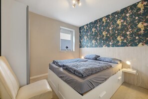 Ein Schlafzimmer mit einem Bett, einem Sessel, einem Nachttisch mit Lampe, einem Fenster mit Jalousien und floralem Wandpapier.
