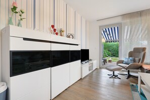 Ein modernes Wohnzimmer mit einem weißen Schrank, einem Fernseher, einem Sessel und Blick auf einen Garten.