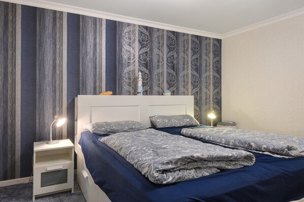A bedroom with two beds, a nightstand with a lamp, and a patterned wallpaper. - Apartment 'Eggers' with Private Terrace, Shared Garden and Wi-Fi (Cuxhaven)