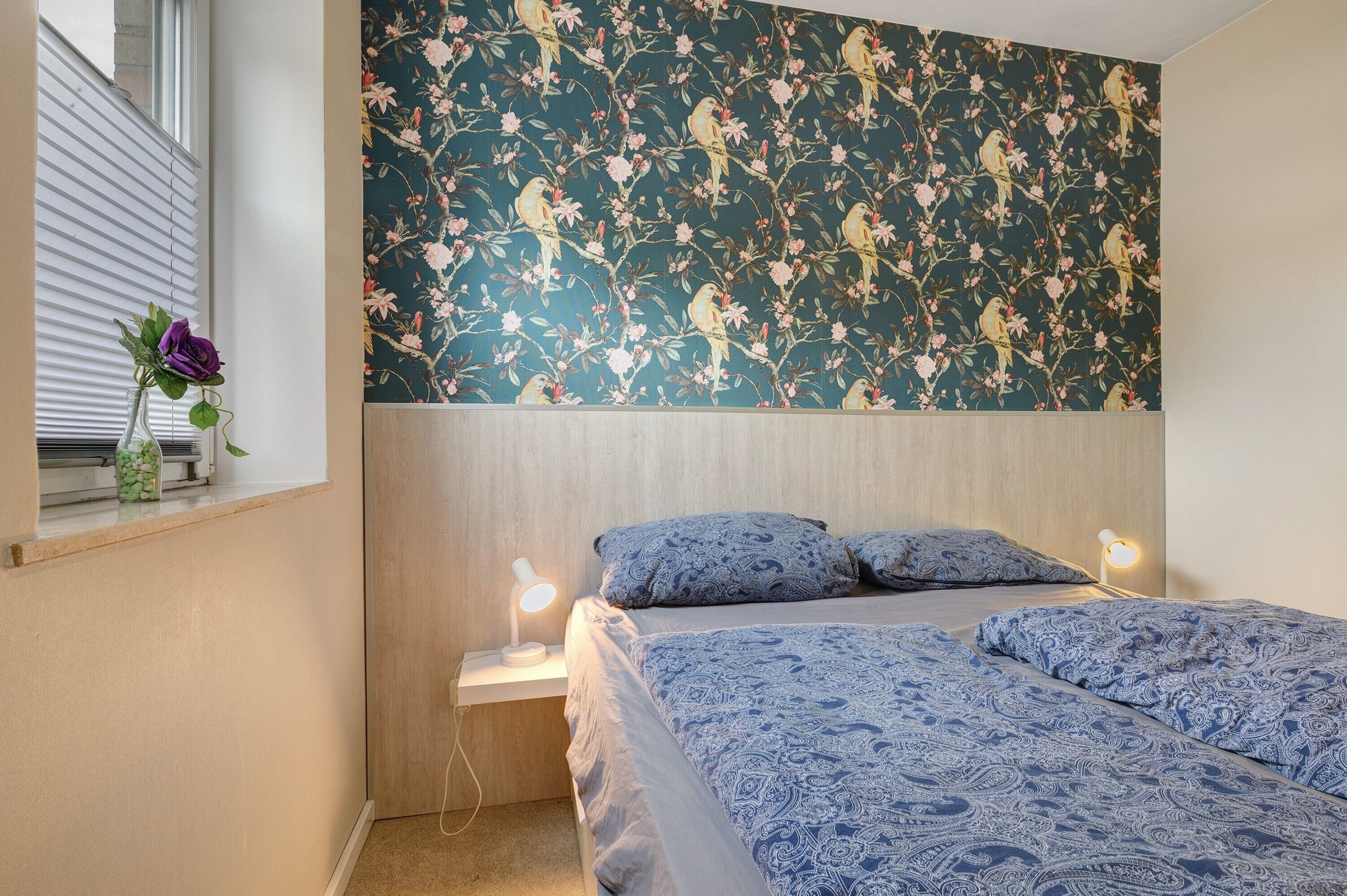 Ein Schlafzimmer mit blumengemustertem Wandpapier, einem Bett mit geblümter Bettwäsche, einem Nachttisch mit Lampe und einem Fenster mit Topfpflanze.