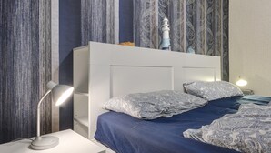 A bedroom with a bed, pillows, a nightstand, a desk lamp, and a wall with vertical patterned wallpaper.