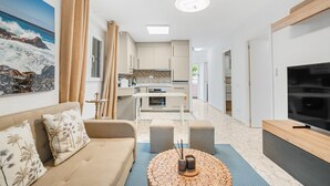 Uma sala de estar moderna com um sofá, uma mesa de centro, uma televisão e uma cozinha ao fundo.