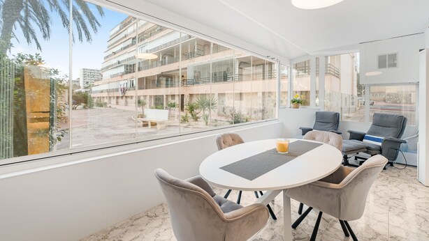 Um interior moderno com uma mesa redonda, cadeiras e uma janela ampla que oferece vista de um edifício de vários andares e área externa.