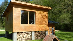 Una cabaña de madera con un ventanal grande, un porche pequeño y una pila de leña.