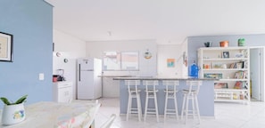 A modern kitchen with a central island, bar stools, a refrigerator, and a bookshelf. - Apartamento em Bertioga com vista para o mar (Bertioga)