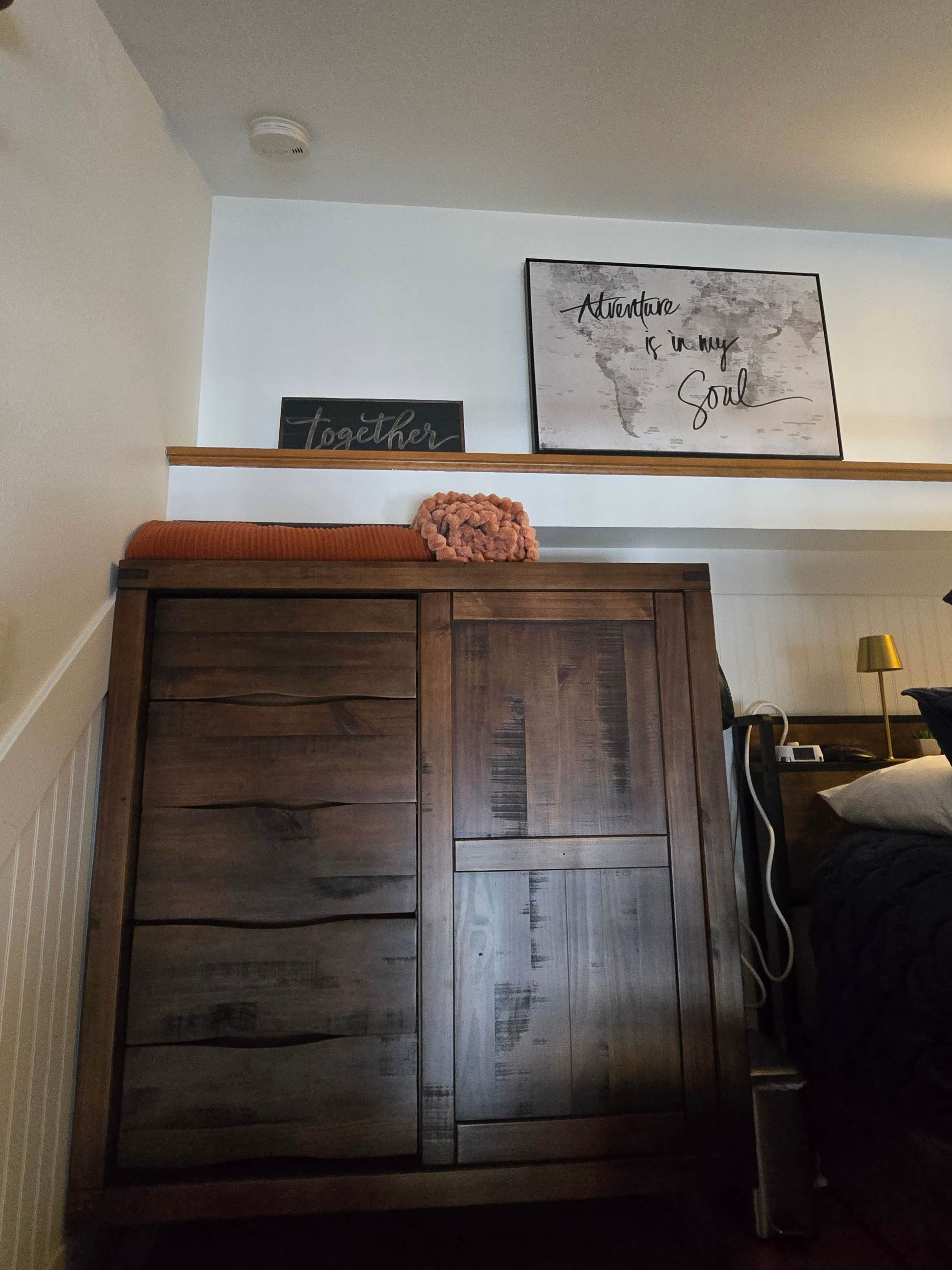 Una camera da letto con un armadio in legno, un letto con cuscino bianco, un comodino con una lampada e una parete decorata con opere d'arte.