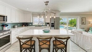 A spacious kitchen with white cabinetry, a central island with bar stools, a stainless steel refrigerator, and a view of a swimming pool.
