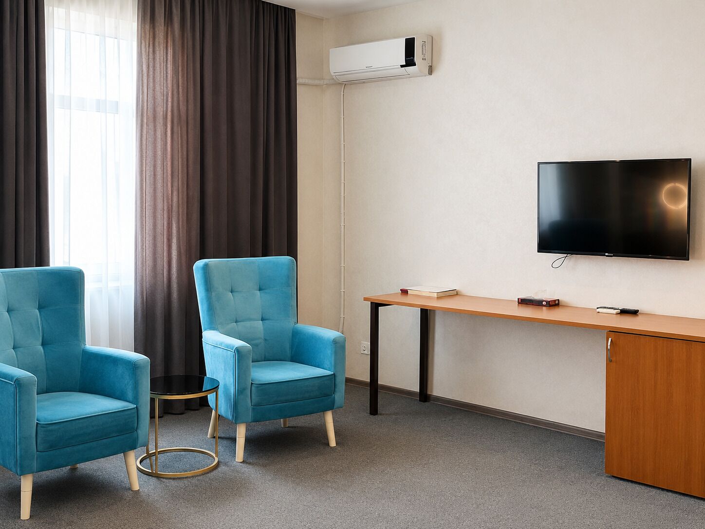 A room with two blue armchairs, a wooden desk, a flat-screen TV, and an air conditioning unit on the wall.