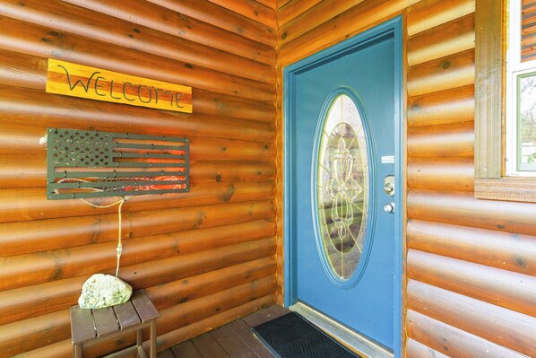 A wooden cabin with a blue door, a welcome sign, and a decorative American flag.