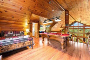 Un interior de madera con mesa de billar, cama con cubrecama escocés y un amplio ventanal con vistas al exterior.