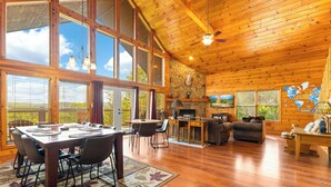 A spacious wooden cabin with a dining area, kitchen, and living room, featuring large windows and a stone fireplace. - 3BDR+Loft for 12 w/Hot Tub, Billiards & Mtn Views! (Sevierville)
