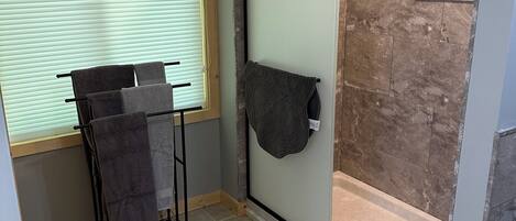 A bathroom with a shower, a towel rack, and a window with blinds.