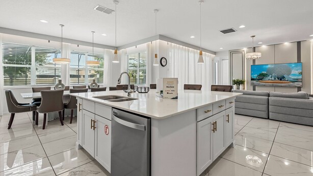 A modern kitchen with a central island, a dining area with chairs, a built-in oven, and a large flat-screen TV.