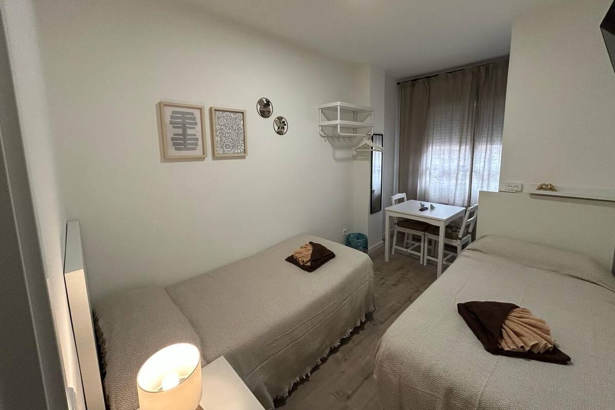 Een hotelkamer met twee bedden, een bureau en een stoel. Er hangen prenten aan de muur, een plafondlamp en een raam met gordijnen.