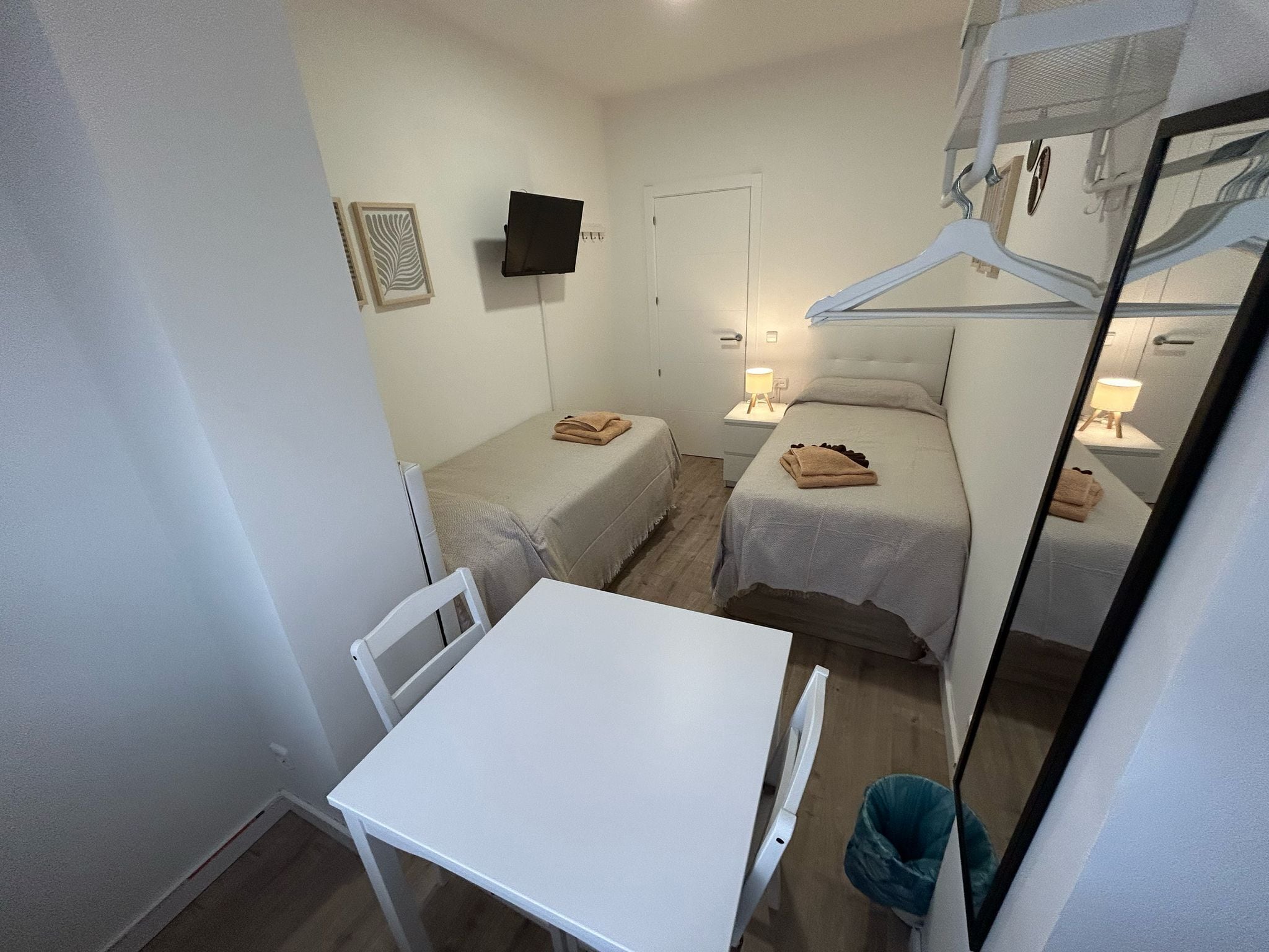 A small, compact room with two single beds, a white table, a chair, a TV mounted on the wall, and a mirror reflecting part of the room.