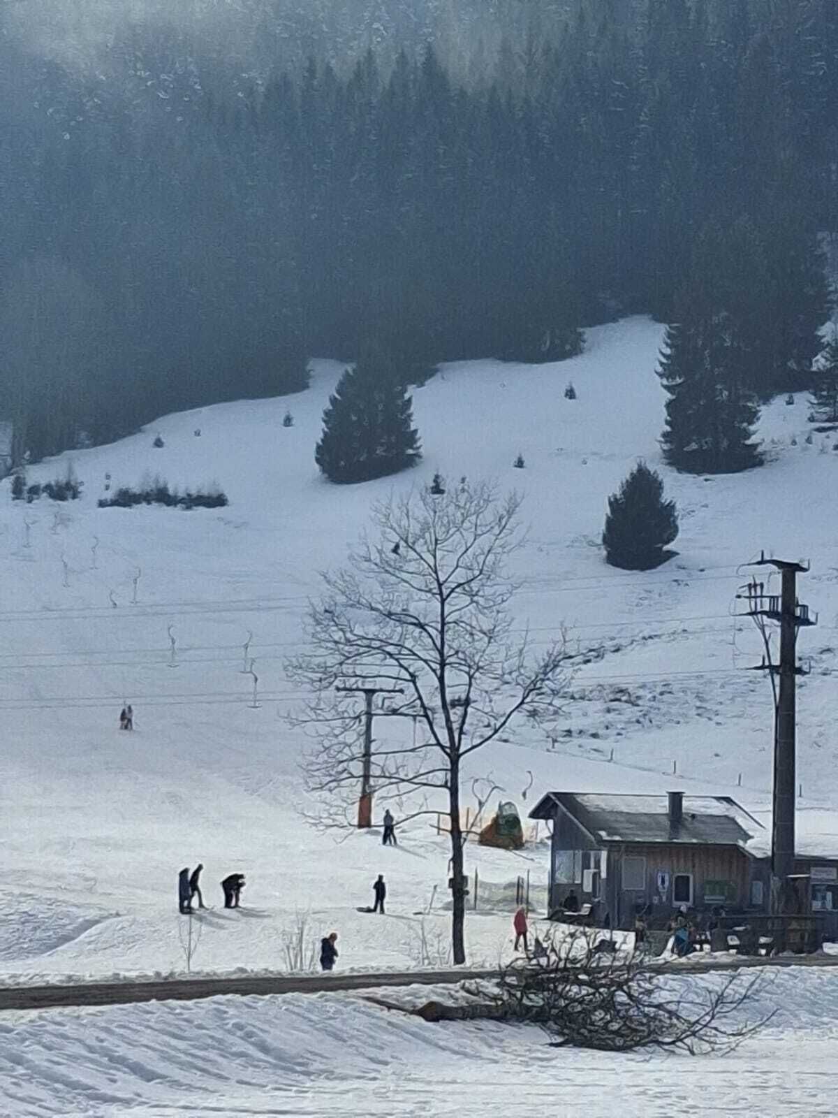 Pendenza innevata per lo sci con alcuni sciatori, una seggiovia e un edificio con tetto spiovente.