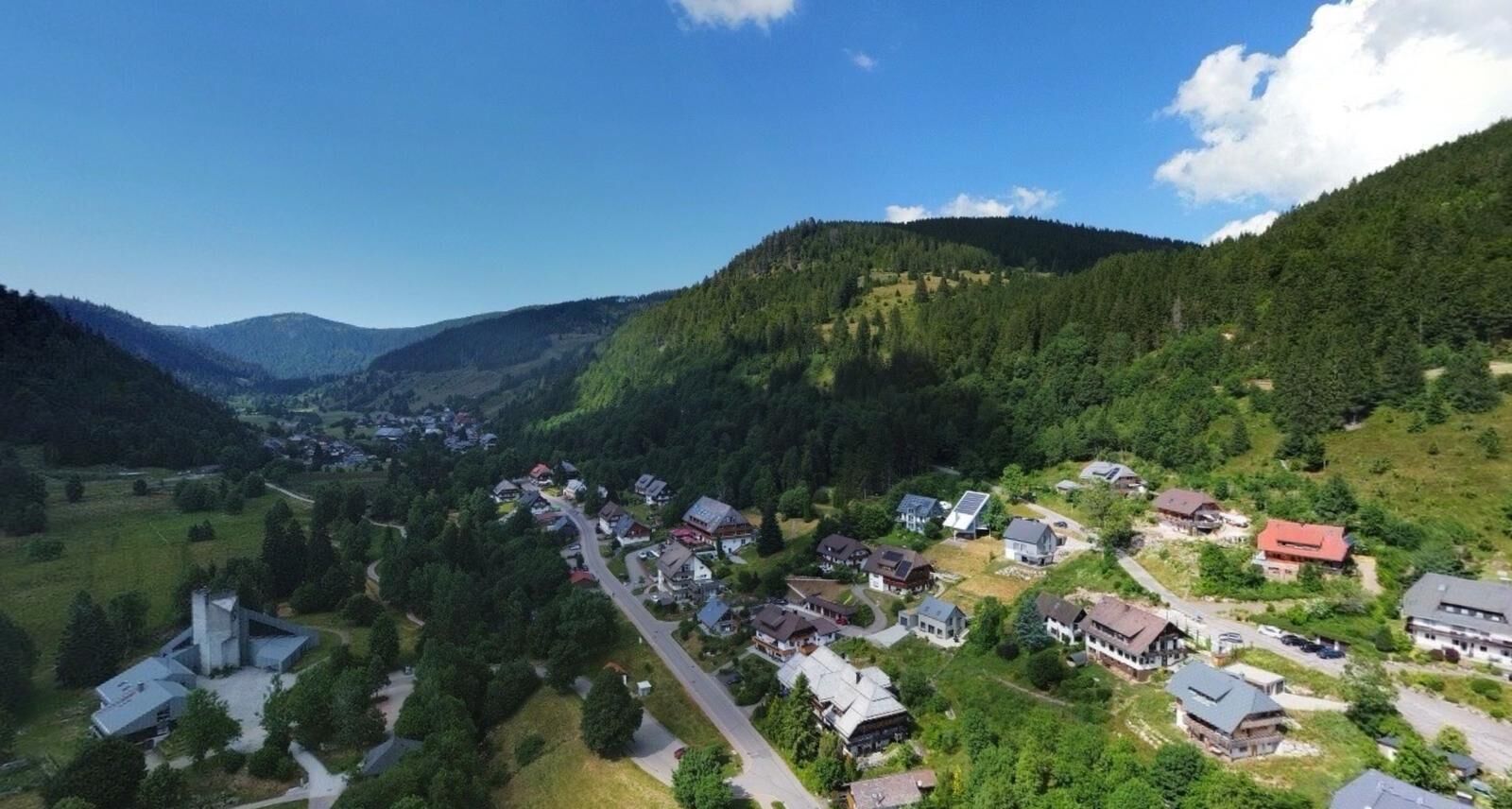 A village with houses surrounded by forested hills, a main road, and a church with a bell tower.