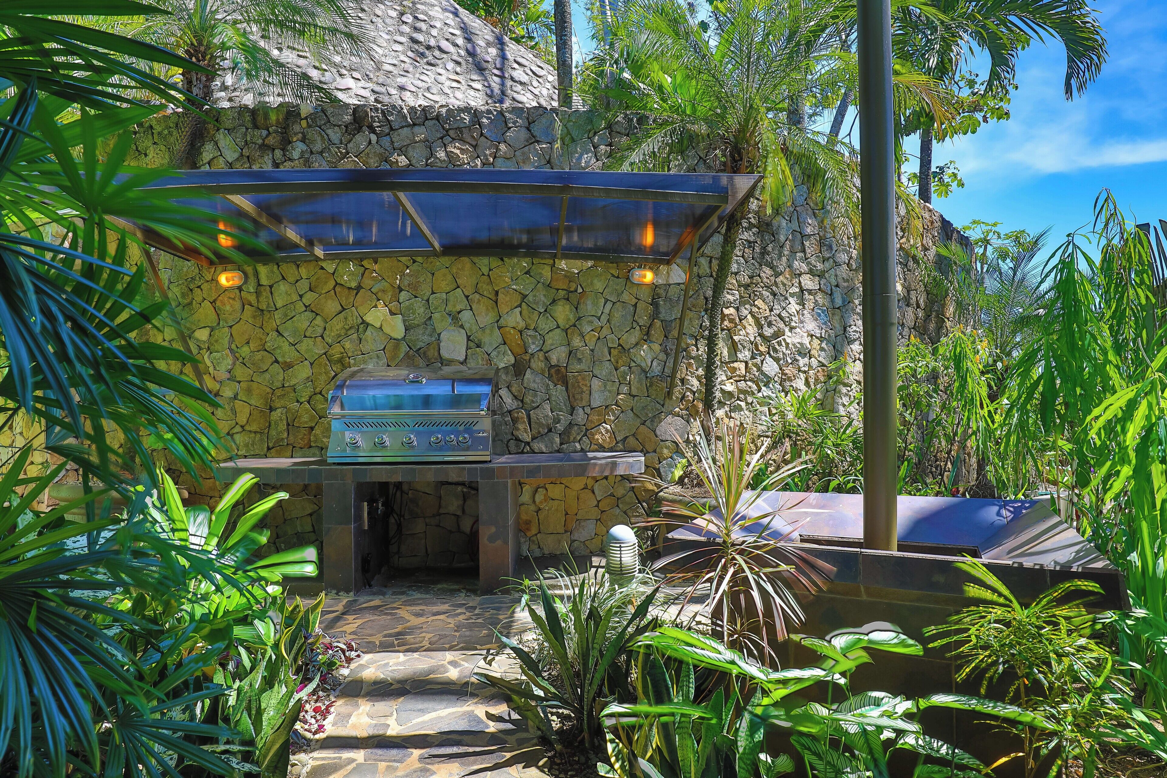 An outdoor barbecue area with a stone wall and grill, surrounded by tropical plants.