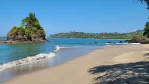 A beach with clear blue water, a sandy shore, and a prominent rock formation with trees. - Casa Pura Vista - Best View Home in Manuel Antonio (Manuel Antonio)