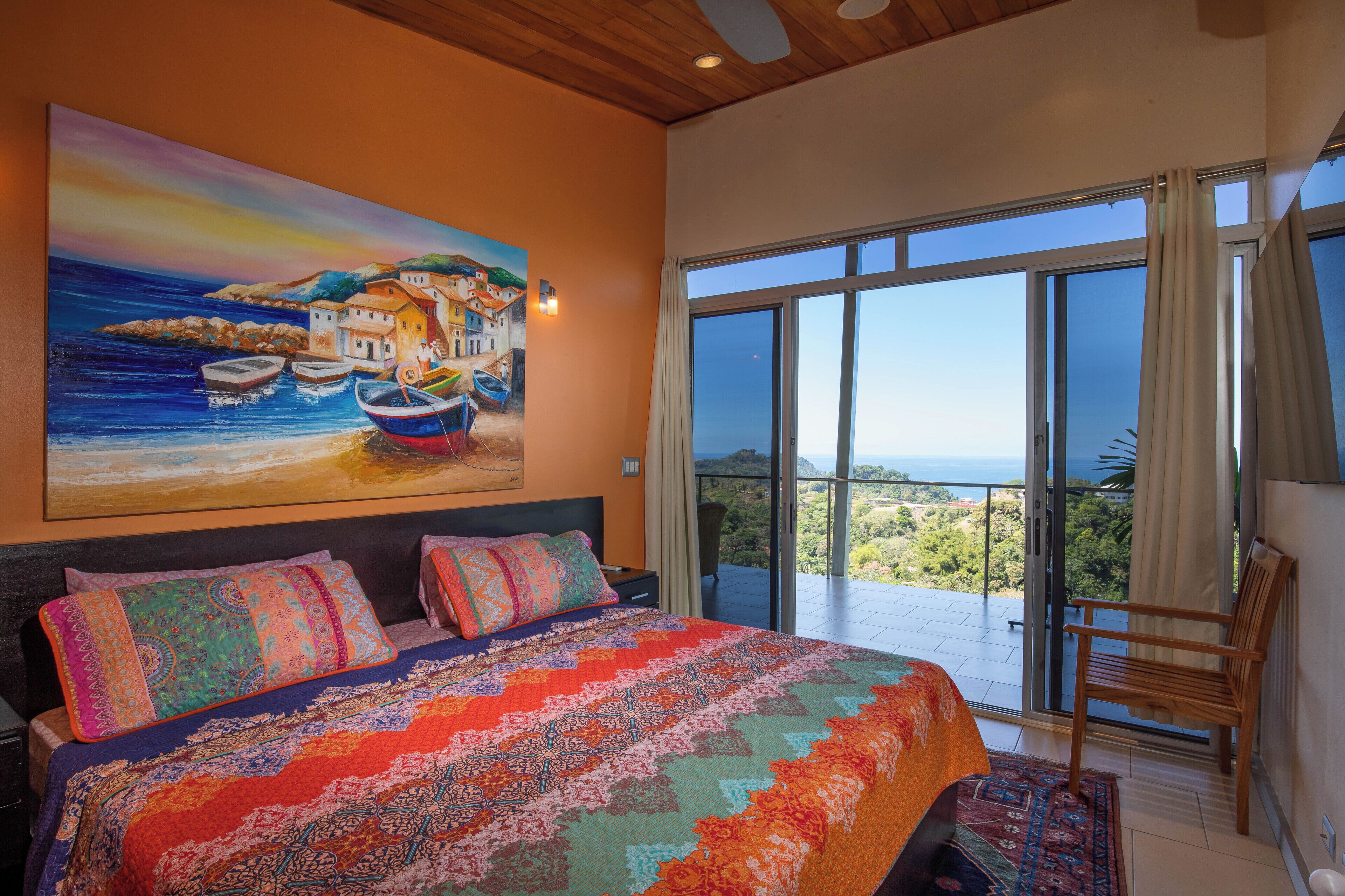 A bedroom with a large bed, colorful bedding, a painting of boats on a cliff, and a balcony with a view.