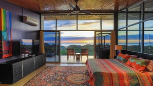 A bedroom with a large bed, a TV, a ceiling fan, and a view of the sunset through large windows. - Casa Pura Vista - Best View Home in Manuel Antonio (Manuel Antonio)