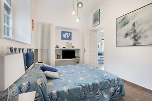 A bedroom with a bed, a radiator, a shelf with books, a TV, and a painting on the wall.