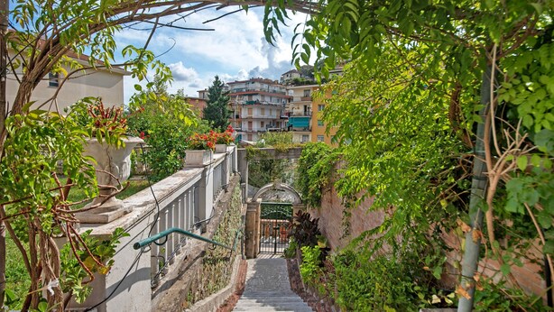 Un escalier menant à une entrée fermée par un portail, entourée de verdure et de bâtiments.