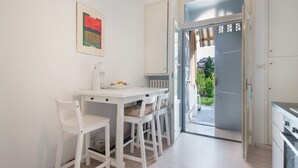 Une cuisine avec des meubles blancs, une table à manger avec des chaises, et une vue sur un balcon, accessible par une porte ouverte.