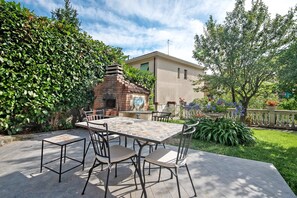 An outdoor patio with a tiled table and chairs, a brick fireplace, and a house in the background.
