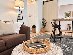 A living room with a brown sofa, a round coffee table with a notebook and pen, a floor lamp, and a dining area with a wooden table and chairs.