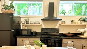 A modern kitchen with a black refrigerator, microwave, stove, and oven. There are two windows with greenery outside, a wooden table set for dining, and various kitchen items on shelves.