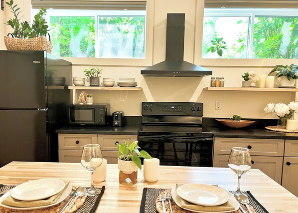 A modern kitchen with a black refrigerator, microwave, stove, and oven. There are two windows with greenery outside, a wooden table set for dining, and various kitchen items on shelves.