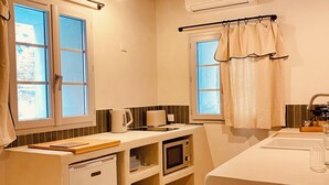 A compact kitchen with a microwave, a small refrigerator, and a sink. There are two windows with curtains, and the ceiling has exposed wooden beams.