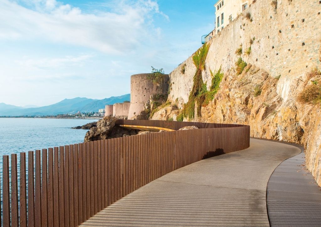 Une promenade en bord de mer, agrémentée d’une clôture en bois, d’un mur de pierre et d’un bâtiment situé sur une falaise surplombant la mer.