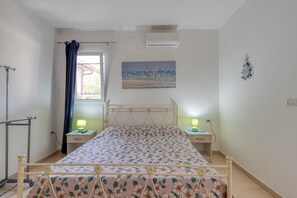Une chambre à coucher comprenant un lit, une table de chevet, une lampe, une fenêtre avec des rideaux, un climatiseur et une horloge murale.