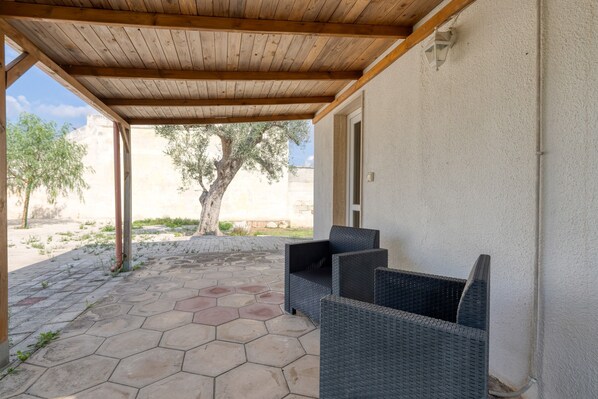 Un patio cubierto con suelo de piedra hexagonal, muebles de mimbre y vista a un árbol y un edificio al fondo.