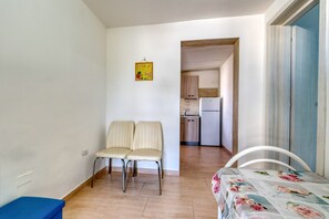 Une petite pièce bien éclairée, avec un lit, deux chaises, une petite table et un coin cuisine au fond.