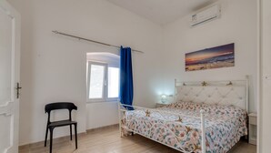 Ein Schlafzimmer mit Bett, Stuhl, Fenster mit Vorhängen, einem Gemälde und einer Klimaanlage.