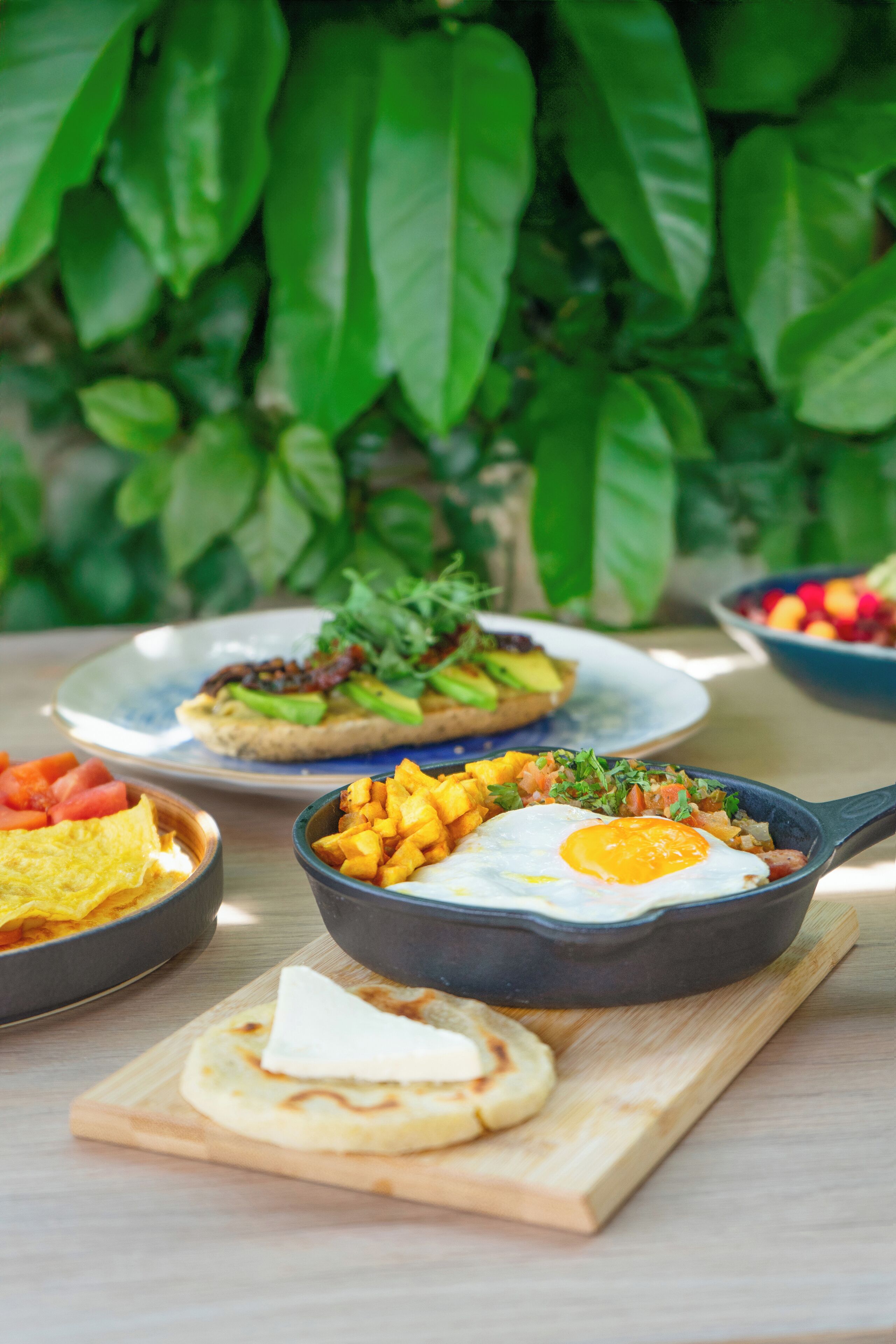 Un desayuno abundante con diversos platos sobre una mesa de madera, rodeado de follaje verde.