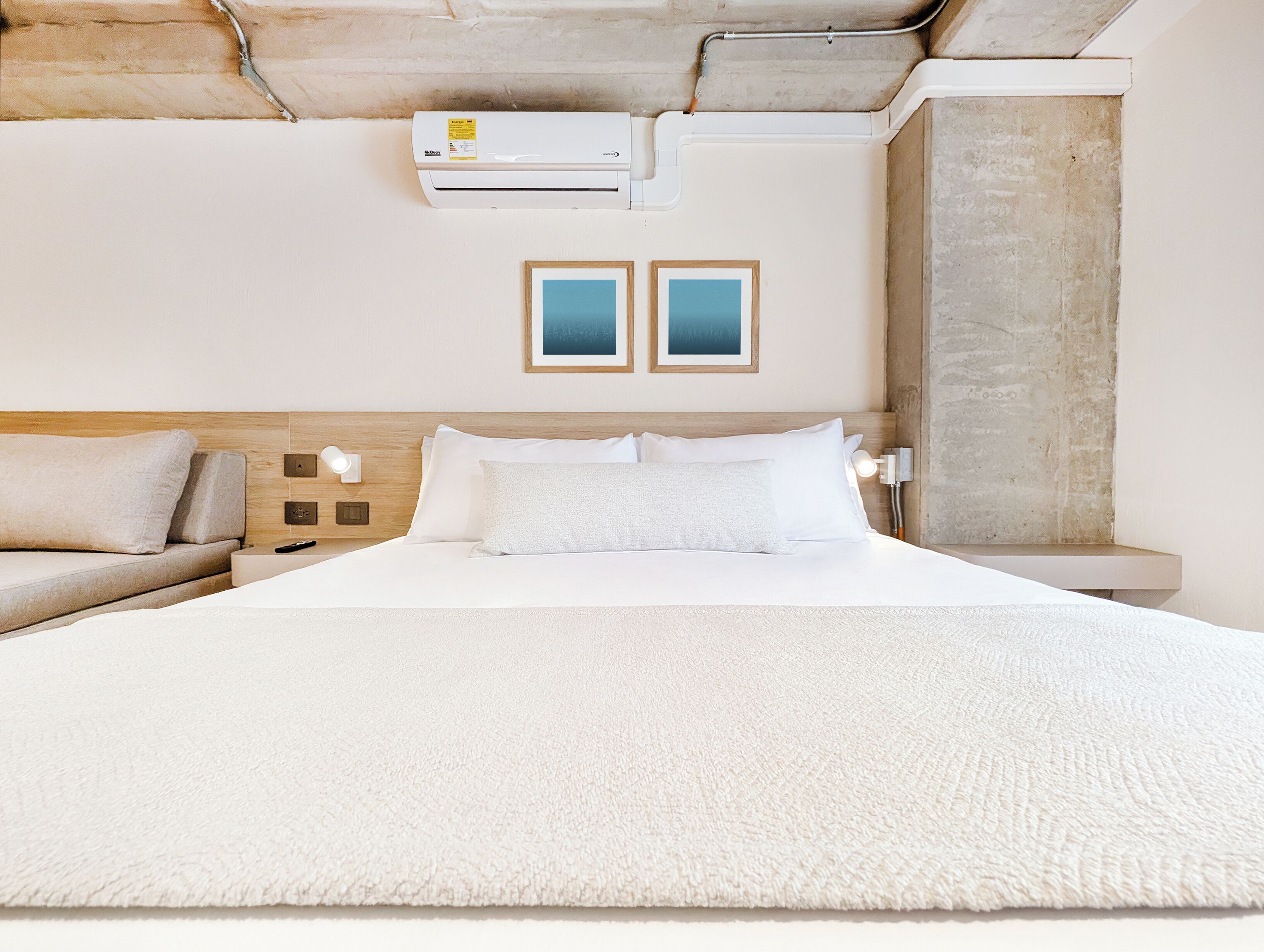 A hotel room with a large bed, a sofa, two wall-mounted picture frames, and an air conditioning unit.