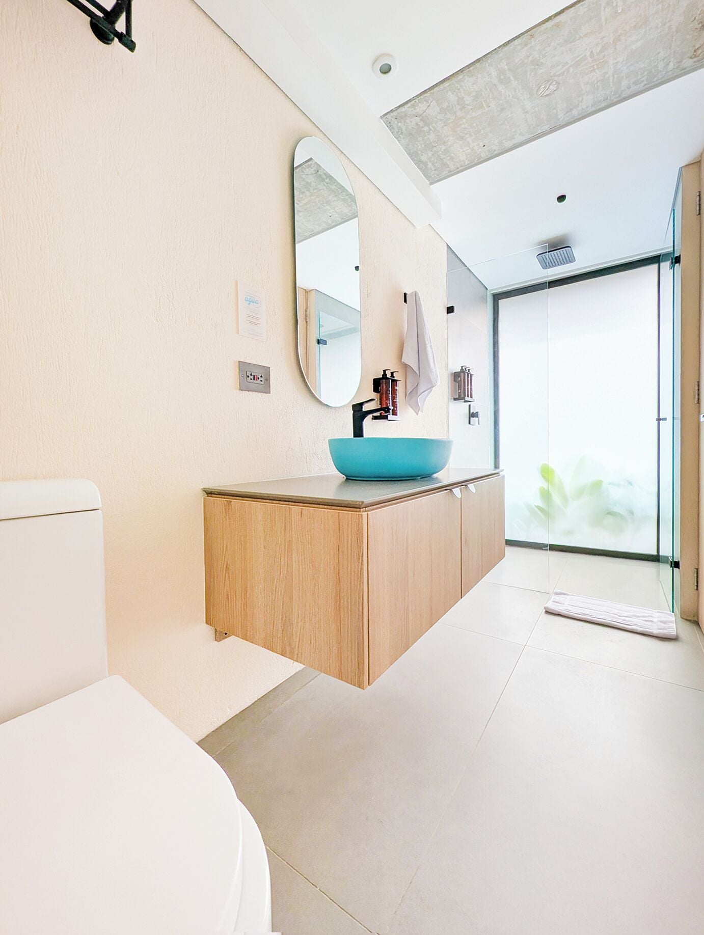 Un baño moderno con inodoro blanco, tocador de madera con lavabo azul, un espejo grande y puerta de ducha de vidrio.